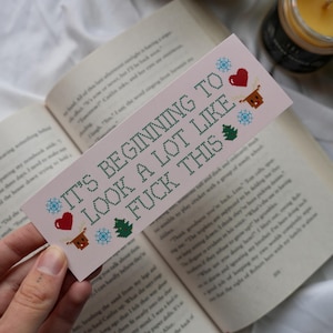 Könnte beinhalten: Ein rosa Lesezeichen mit dem Text "IT'S BEGINNING TO LOOK A LOT LIKE FUCK THIS" in Grün, mit pixeligen Schneeflocken, einem Herz, einem Rentier und einem Weihnachtsbaum. Das Lesezeichen liegt auf einem offenen Buch.