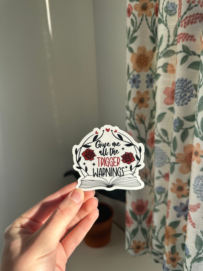 May include: A white sticker with a red open book design. The book has a red rose on each side and the text "Give me all the trigger warnings" in black.