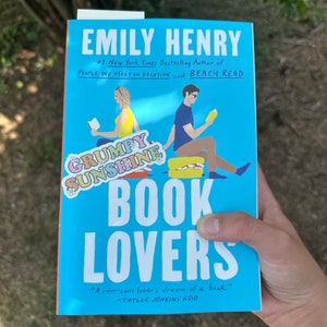 May include: A blue book cover with the title "Book Lovers" and the author's name "Emily Henry". The cover features an illustration of two people sitting on a bench reading books. The text "#1 New York Times Bestselling Author of People We Meet on Vacation and Beach Read" is also on the cover.