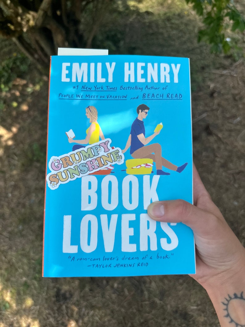 May include: A blue book cover with the title "Book Lovers" and the author's name "Emily Henry". The cover features an illustration of two people sitting on a bench reading books. The text "#1 New York Times Bestselling Author of People We Meet on Vacation and Beach Read" is also on the cover.