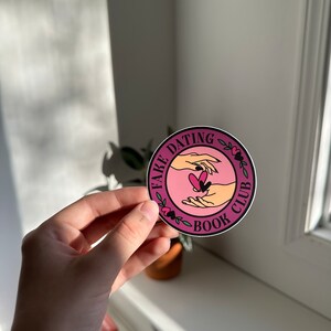 May include: A pink and white round sticker with the text "Fake Dating Book Club" and a graphic of two hands holding a heart.