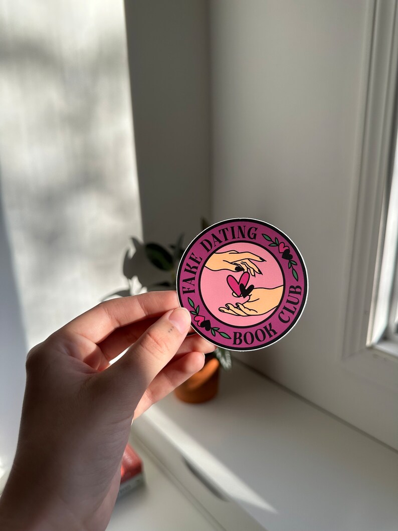 May include: A pink and white round sticker with the text "Fake Dating Book Club" and a graphic of two hands holding a heart.