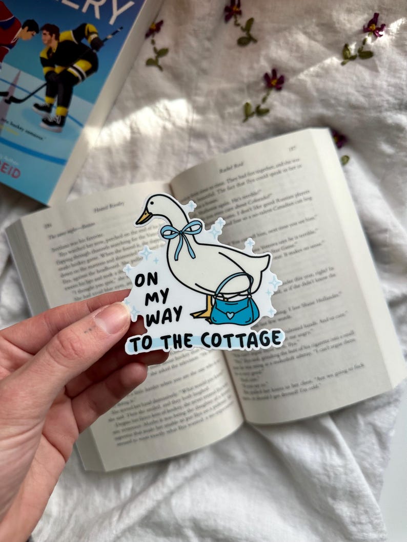 May include: A white sticker featuring a cartoon goose with a blue bow and purse. Text on the sticker reads "ON MY WAY TO THE COTTAGE". The sticker is held over an open book with a book cover in the background.