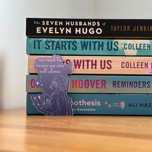May include: A stack of books with a purple and white sticker of a person reading. The sticker says "we read to know we are not alone". The book titles are "The Seven Husbands of Evelyn Hugo", "It Starts With Us", "Reminders", and "The Hypothesis".