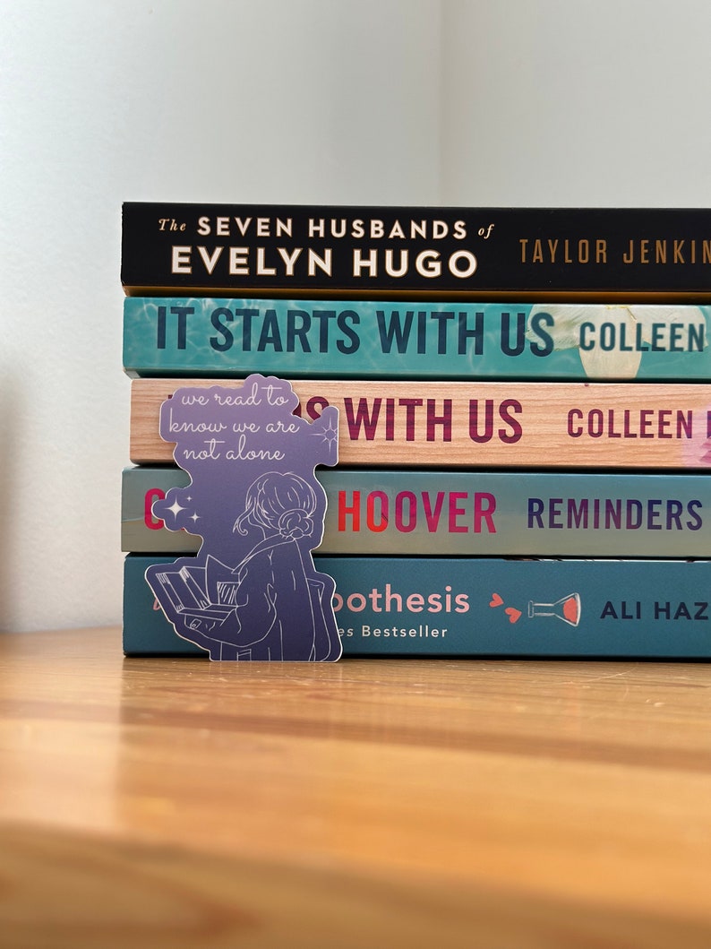 May include: A stack of books with a purple and white sticker of a person reading. The sticker says "we read to know we are not alone". The book titles are "The Seven Husbands of Evelyn Hugo", "It Starts With Us", "Reminders", and "The Hypothesis".