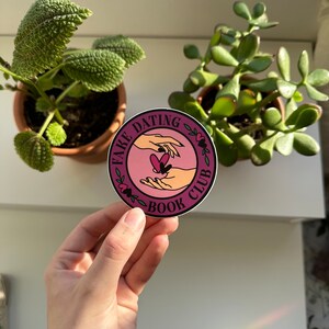 May include: A pink and white round sticker with the text "Fake Dating Book Club" in a circle with a floral design. Two hands are reaching towards each other with a heart in between them.