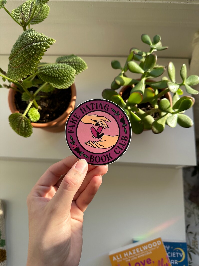 May include: A pink and white round sticker with the text "Fake Dating Book Club" in a circle with a floral design. Two hands are reaching towards each other with a heart in between them.