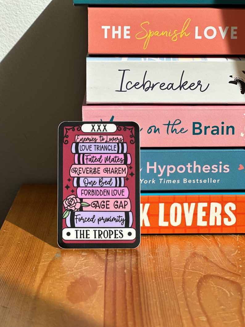 May include: A sticker with a black border and a pink and purple background. The sticker features a stack of books with the text "XXX" at the top and "THE TROPES" at the bottom. The books are labeled with common romance tropes, including "Enemies to Lovers", "Love Triangle", "Fated Mates", "Reverse Harem", "One Bed", "Forbidden Love", "Age Gap", and "Forced Proximity".