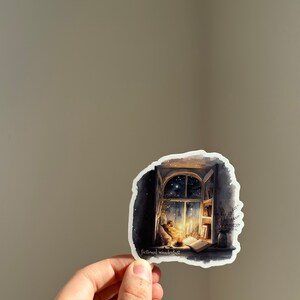 以下が含まれることがあります： 星空の夜を望む窓を描いた水彩画のステッカー。窓際で本を読み、ろうそくを灯している人がいます。ステッカーの下部には「fictional wanderlust」の文字があります。