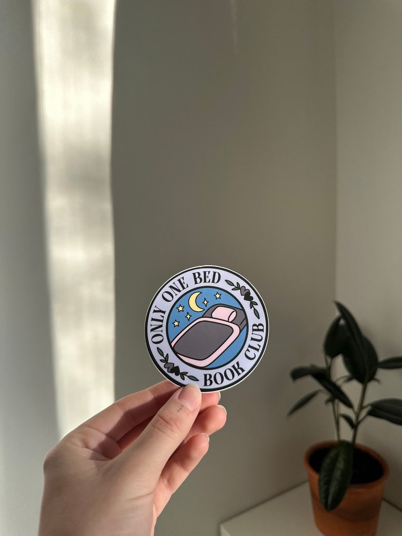 May include: A round sticker with a blue background and white text that reads "Only One Bed Book Club". The sticker features a cartoon illustration of a bed with a pillow and blanket, a crescent moon, and stars.