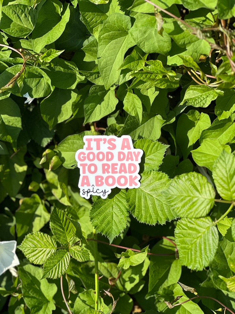 May include: A pink sticker with the text "It's a good day to read a book" with the word "spicy" written underneath in a smaller font. The sticker is placed on a bed of green leaves.