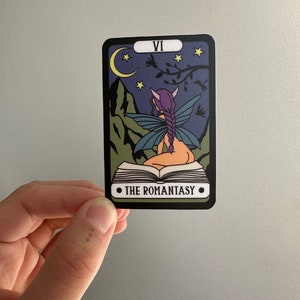 May include: A tarot card with a purple fairy sitting on a book. The card is black with a white border and the number VI and the text "THE ROMANTASY" are printed on the card. The background is a dark blue night sky with a crescent moon and stars.