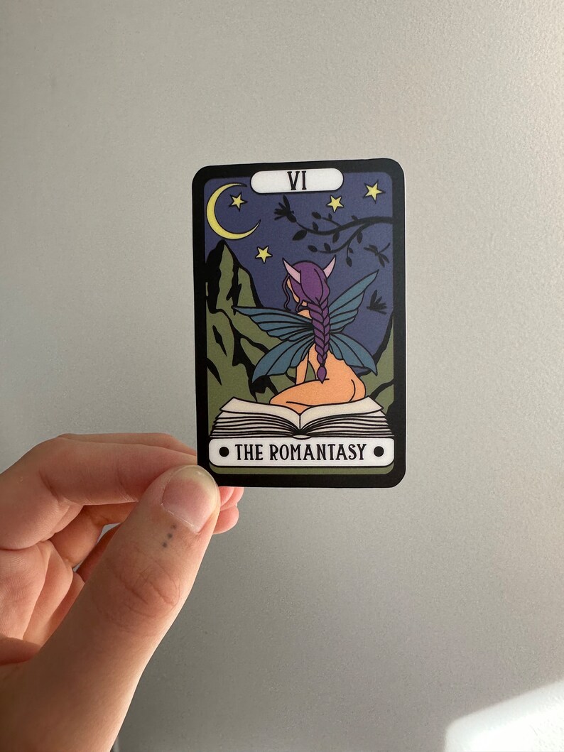 May include: A tarot card with a purple fairy sitting on a book. The card is black with a white border and the number VI and the text "THE ROMANTASY" are printed on the card. The background is a dark blue night sky with a crescent moon and stars.