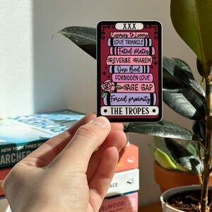 May include: A sticker with a red background and a stack of books with different romance tropes written on them. The text on the sticker reads "XXX Enemies to Lovers Love Triangle Fated Mates Reverse Harem One Bed Forbidden Love Age Gap Forced Proximity The Tropes".