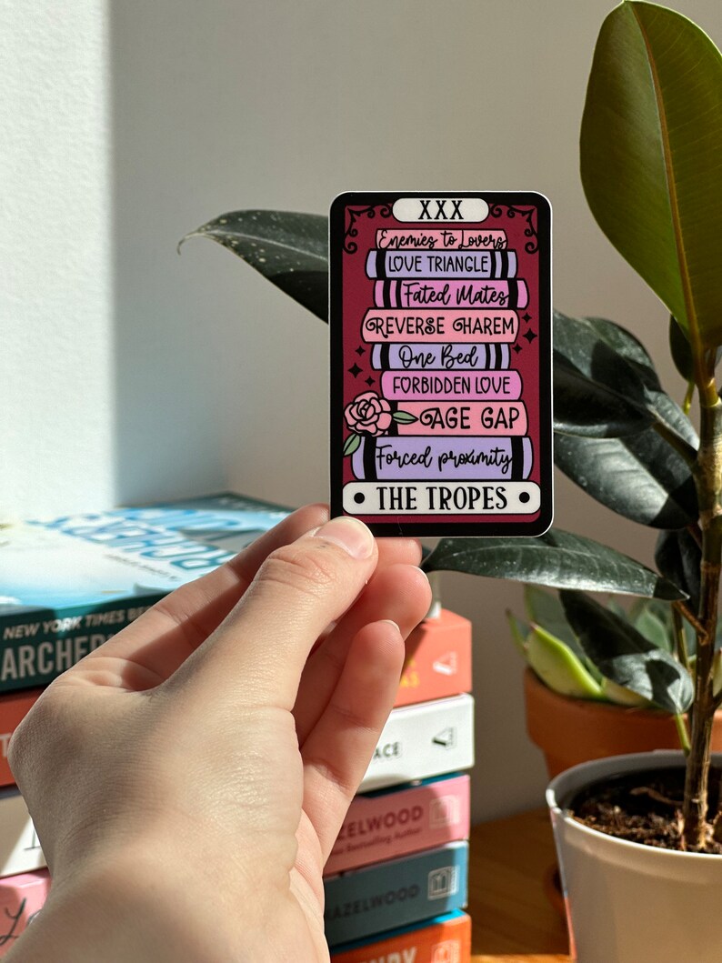 May include: A sticker with a red background and a stack of books with different romance tropes written on them. The text on the sticker reads "XXX Enemies to Lovers Love Triangle Fated Mates Reverse Harem One Bed Forbidden Love Age Gap Forced Proximity The Tropes".