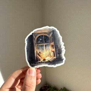 以下が含まれることがあります： 星空の夜空が見える窓を描いた水彩画のステッカーです。窓が開いており、中にはテーブルの上に本とろうそくがあります。ステッカーの下部には「fictional wanderlust」というテキストが印刷されています。