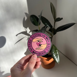 May include: A pink and white round sticker with the text "Fake Dating Book Club" and a graphic of two hands holding a heart.