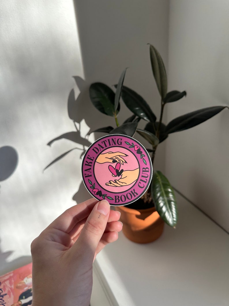 May include: A pink and white round sticker with the text "Fake Dating Book Club" and a graphic of two hands holding a heart.