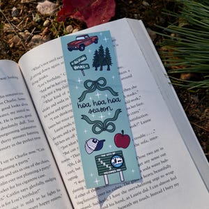 May include: A teal bookmark with various illustrations, including a red truck, pine trees, bows, an apple, and a baseball cap. The text "HOA HOA HOA SEASON" and "The City of Forks Welcomes You" are also featured.
