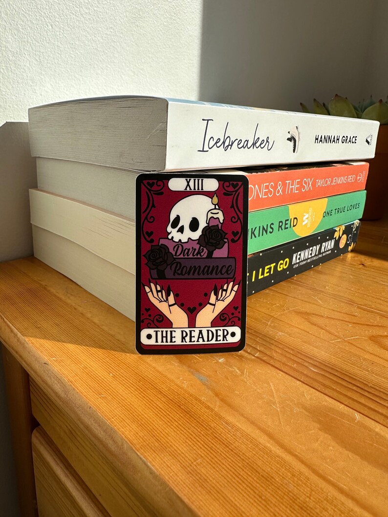 May include: A tarot card with a red background and a skull with a candle on top. The card says "Dark Romance" and "The Reader" in black text. The card is numbered "XIII".
