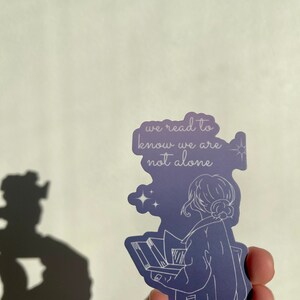 May include: A purple sticker with a white outline of a person reading a book. The text on the sticker reads "we read to know we are not alone".