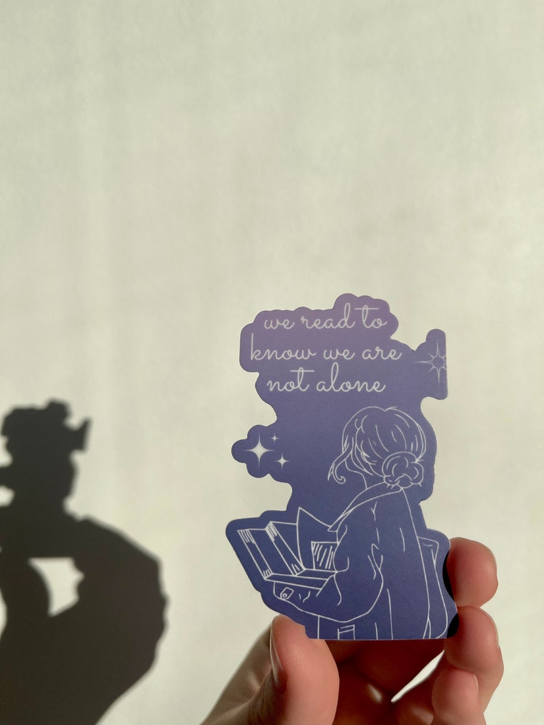 May include: A purple sticker with a white outline of a person reading a book. The text on the sticker reads "we read to know we are not alone".