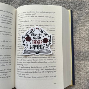 May include: A close-up of a book with a sticker on the page. The sticker is a white circle with a red rose and the text "Give me all the trigger warnings". The book is open to a page with text about a floating island and a girl who is able to save the world from a power-hungry man's evil ambitions.