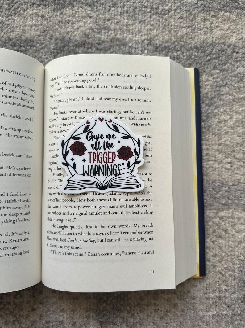 May include: A close-up of a book with a sticker on the page. The sticker is a white circle with a red rose and the text "Give me all the trigger warnings". The book is open to a page with text about a floating island and a girl who is able to save the world from a power-hungry man's evil ambitions.