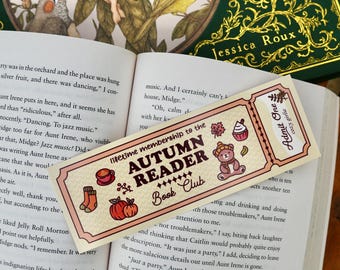 May include: A bookmark with the text "AUTUMN READER Book Club" and illustrations of fall-themed items like a bear, pumpkin, and cup. The bookmark is on an open book with a green book in the background.