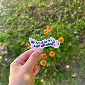 以下が含まれることがあります： ピンクのハート型のステッカーに、黒い文字で「my heart belongs to zane meadows」と書かれています。