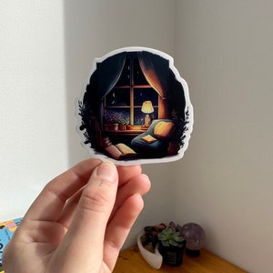 May include: A sticker featuring a cozy nighttime scene. The illustration depicts a window with a view of a starry sky, a lit lamp, and a comfortable armchair. The sticker has a white border and is held by a hand.