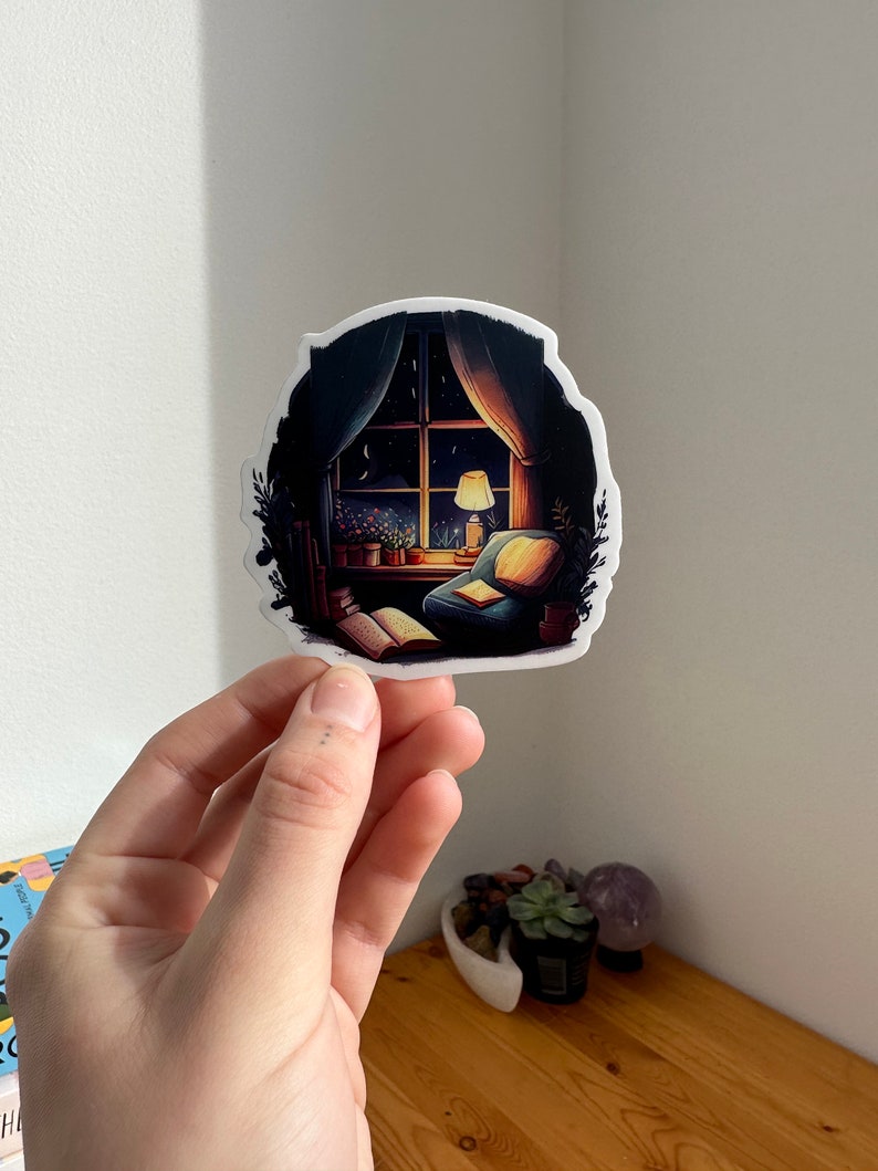 May include: A sticker featuring a cozy nighttime scene. The illustration depicts a window with a view of a starry sky, a lit lamp, and a comfortable armchair. The sticker has a white border and is held by a hand.