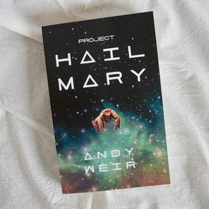 May include: A book titled "Project Hail Mary" by Andy Weir. The cover shows a starry night sky with a nebula and a space-suited figure. The book's title and author's name are in white. The book is on a white, textured surface.