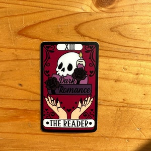 May include: A tarot card with a red background and black and white illustrations. The card is titled "The Reader" and features a skull, a candle, and two hands with long nails. The text "Dark Romance" is also featured on the card.