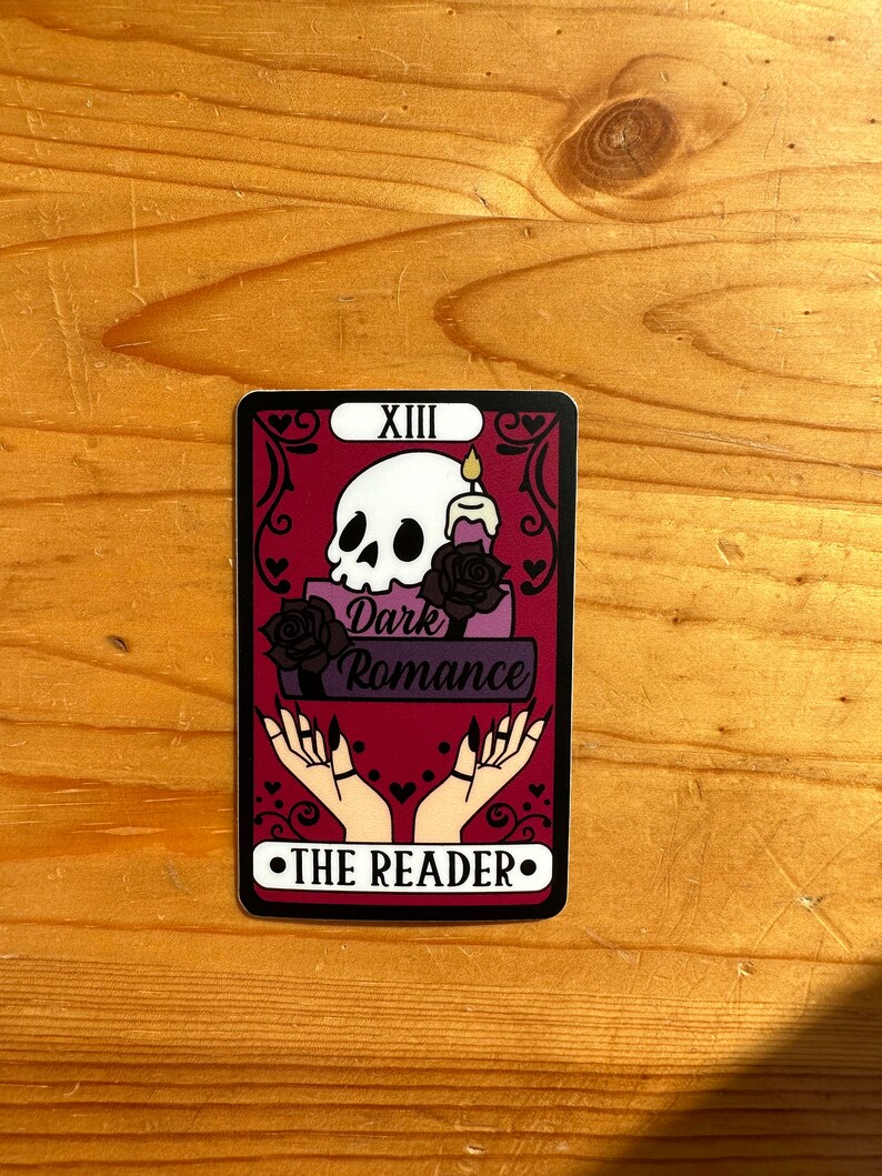 May include: A tarot card with a red background and black and white illustrations. The card is titled "The Reader" and features a skull, a candle, and two hands with long nails. The text "Dark Romance" is also featured on the card.