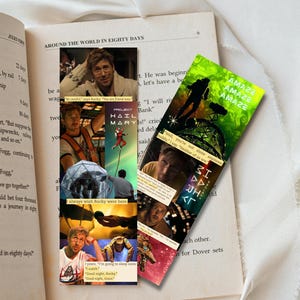 May include: Two colorful bookmarks featuring images from a movie, placed on an open book. One bookmark has a green background with the word "AMAZE" repeated. The other bookmark has images of a man and the text "PROJECT HAIL MARY".