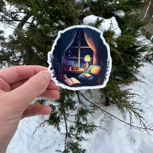 May include: A sticker featuring a cozy illustration of a room with a window overlooking a night sky. Inside, a lit lamp, open book, and armchair create a warm, inviting scene. The sticker is held against a backdrop of snow and evergreen branches.