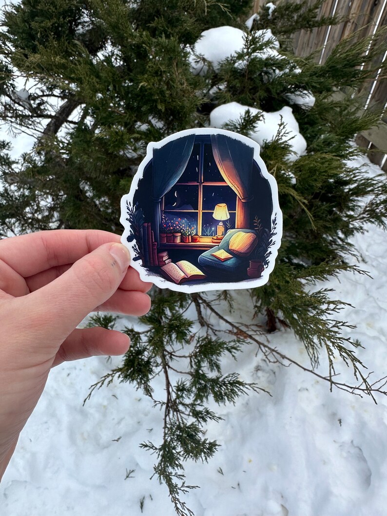 May include: A sticker featuring a cozy illustration of a room with a window overlooking a night sky. Inside, a lit lamp, open book, and armchair create a warm, inviting scene. The sticker is held against a backdrop of snow and evergreen branches.