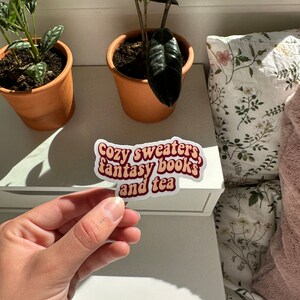 K&ouml;nnte beinhalten: Ein Aufkleber mit einem Retro-Schriftdesign, auf dem "Cozy sweaters, fantasy books, and tea" steht. Der Aufkleber ist braun und wei&szlig; mit einem roten Rand.