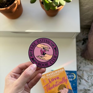 May include: A purple and black round sticker with the text "Fake Dating Book Club" and a graphic of two hands holding a heart.