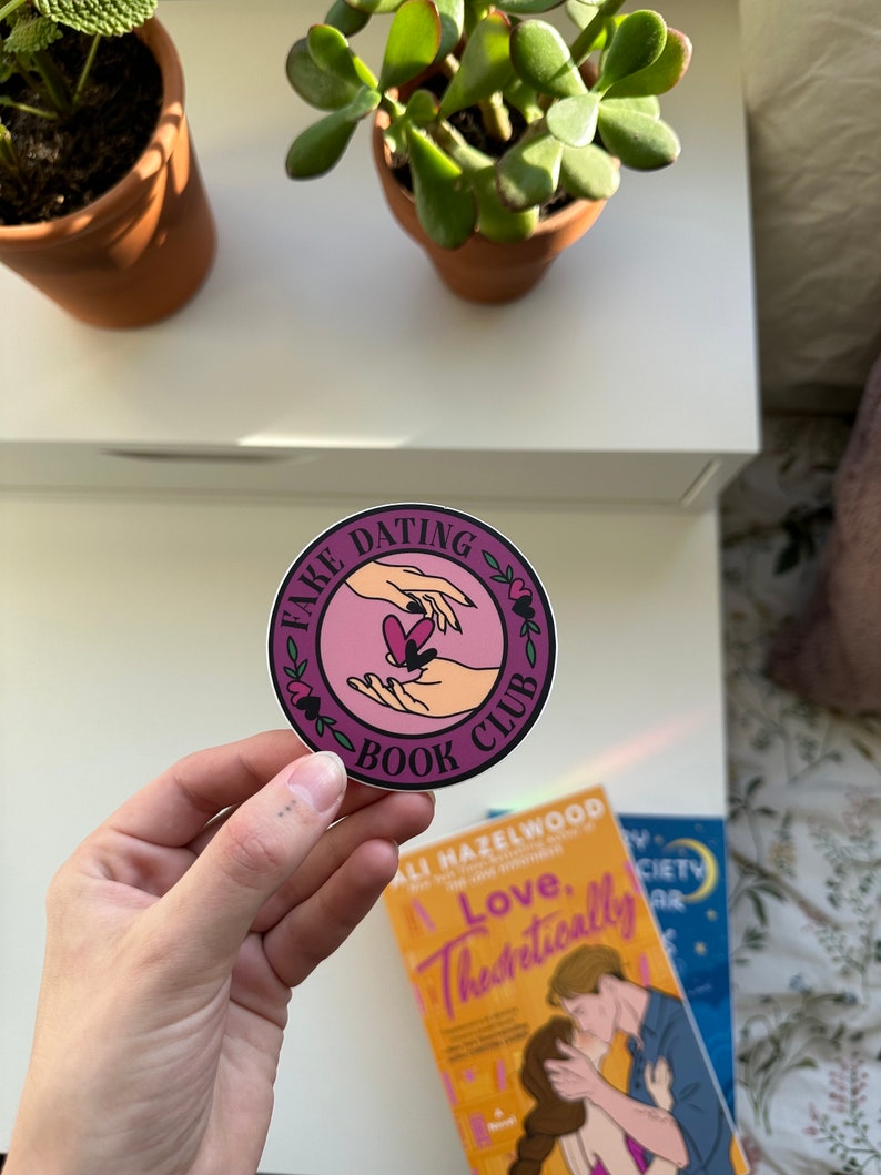 May include: A purple and black round sticker with the text "Fake Dating Book Club" and a graphic of two hands holding a heart.