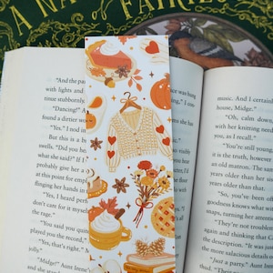May include: A bookmark with autumnal illustrations, including a cream cardigan with red hearts, pumpkins, pie, coffee, and leaves. The bookmark is in an open book titled "A Natural History of Fairies". The book is in English.