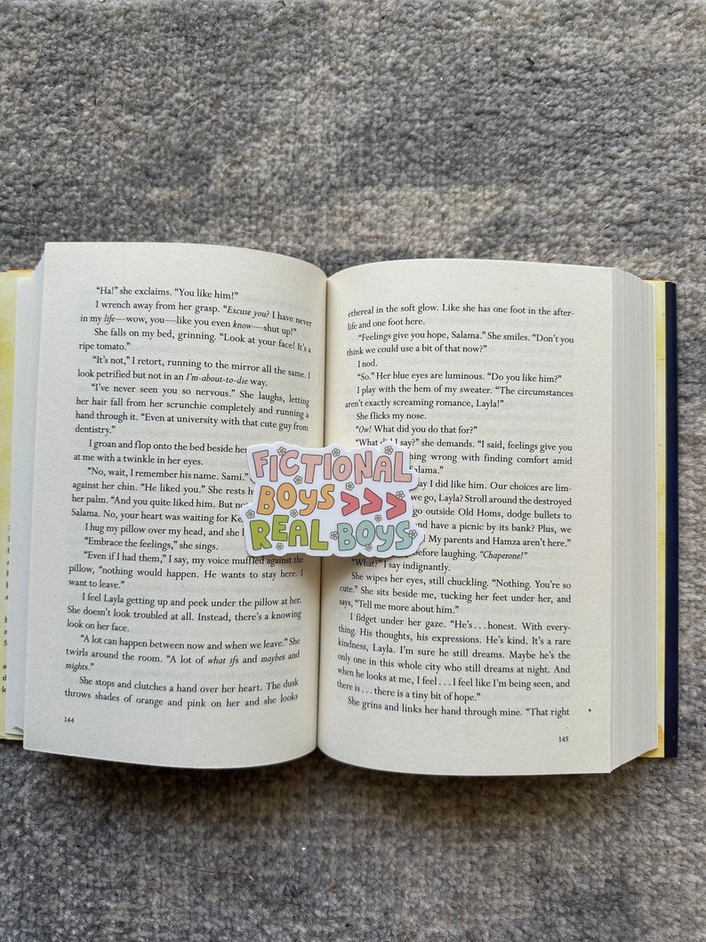 K&ouml;nnte beinhalten: Eine Nahaufnahme eines Buches, das auf einer Seite mit Text &uuml;ber die Gef&uuml;hle einer jungen Frau ge&ouml;ffnet ist. Der Text lautet "Feelings give you hope, Salama." Das Buch liegt auf einem Bett mit einer bunten Tagesdecke.