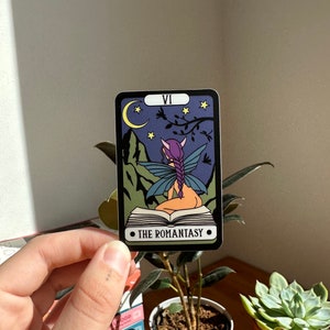 May include: A tarot card with a purple-haired fairy sitting on a book. The card is black with a purple, green, and blue design. The card is labeled "VI" and "THE ROMANTASY".