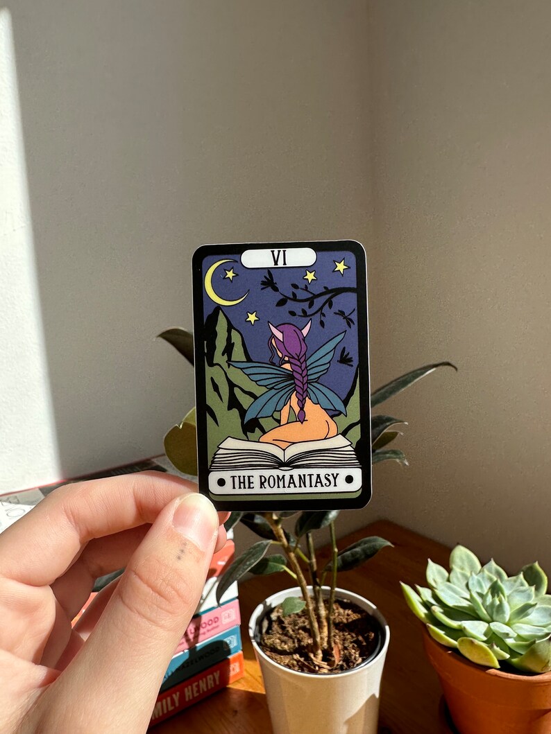 May include: A tarot card with a purple-haired fairy sitting on a book. The card is black with a purple, green, and blue design. The card is labeled "VI" and "THE ROMANTASY".