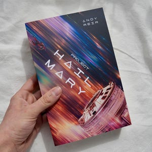 Op de afbeelding: Een hardcover boek met de titel "Project Hail Mary" van Andy Weir. De omslag toont een ruimtescène met een ruimteschip en een kleurrijke, gestreepte achtergrond. Het boek wordt door een persoon vastgehouden.