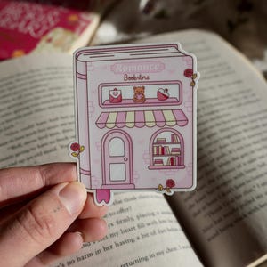 May include: A pink sticker depicting a bookstore, labelled "Romance Bookstore." The sticker shows a door, a window with books, and a shelf with a teddy bear, envelope, and heart. Roses decorate the edges of the sticker.