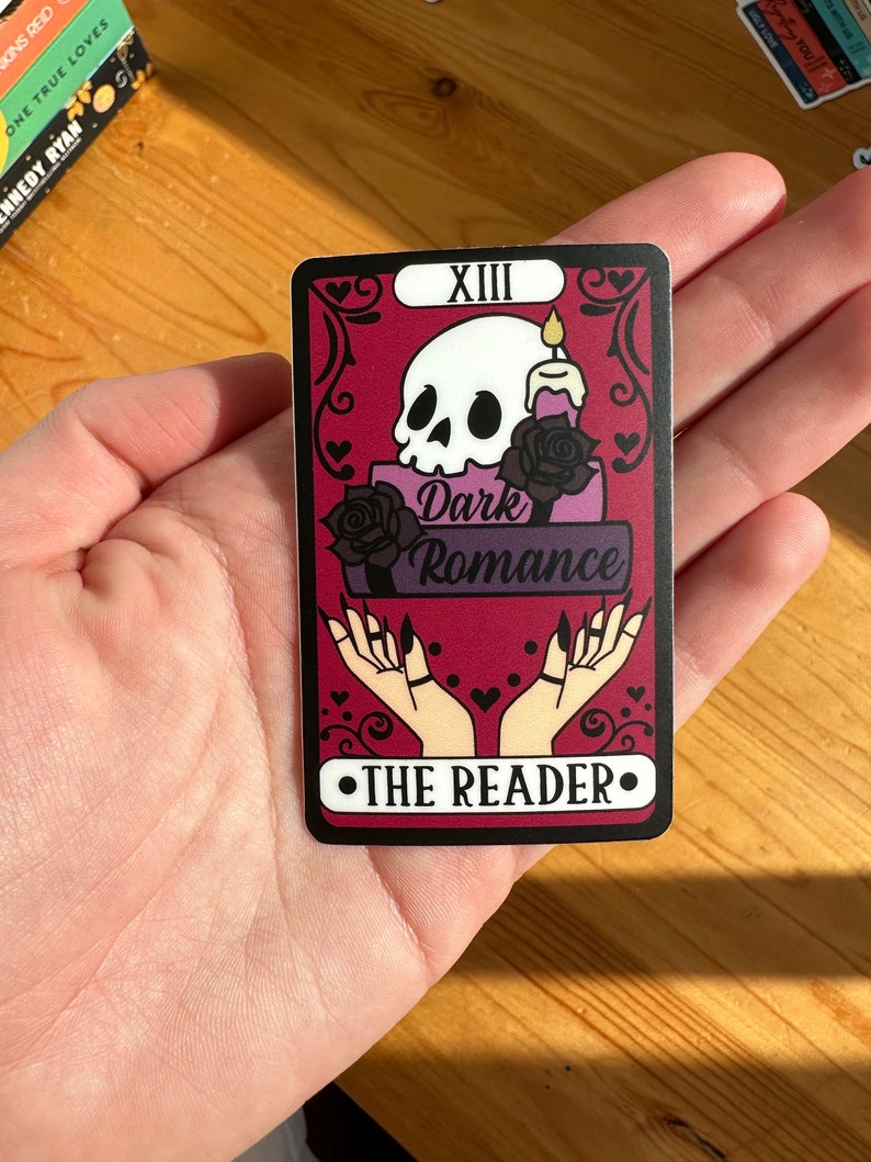May include: A tarot card with a black border and a red background. The card features a skull with a candle on top of it, and the words "Dark Romance" written below. The card is labeled "XIII" and "THE READER" in black text.