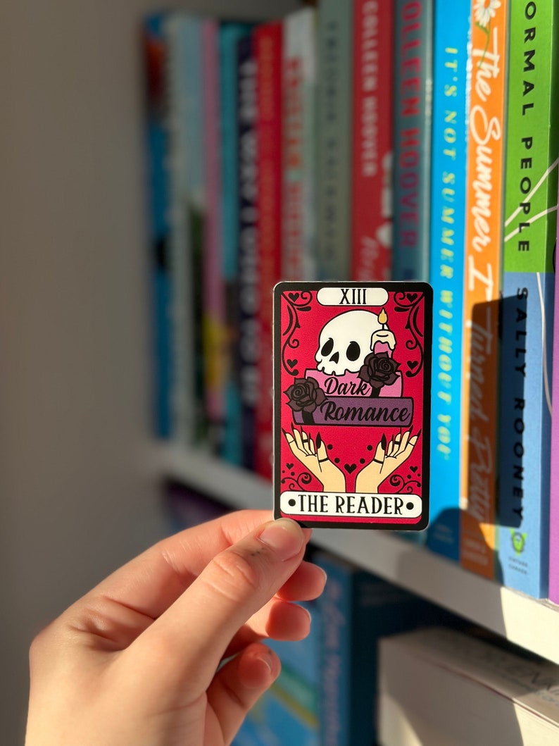 May include: A tarot card with a pink and purple background. The card features a skull with a candle on top of it, a black rose, and two hands with long black nails. The card is labeled "XIII Dark Romance" and "THE READER".