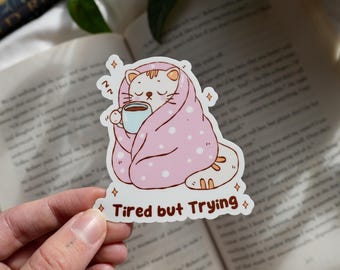 May include: A sticker featuring a cartoon cat wrapped in a pink blanket with white polka dots, holding a blue mug. The text "Tired but Trying" is below the cat. The sticker is held in front of an open book.